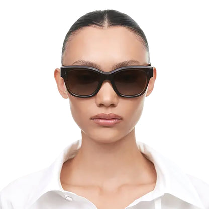 A model with slicked-back hair wearing chunky black cat-eye sunglasses with dark brown lenses, paired with a white collared shirt against a plain white background.