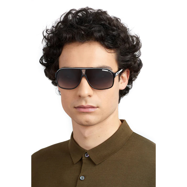 A man with curly dark hair wearing dark aviator-style sunglasses and an olive brown polo shirt, shown against a plain white background.